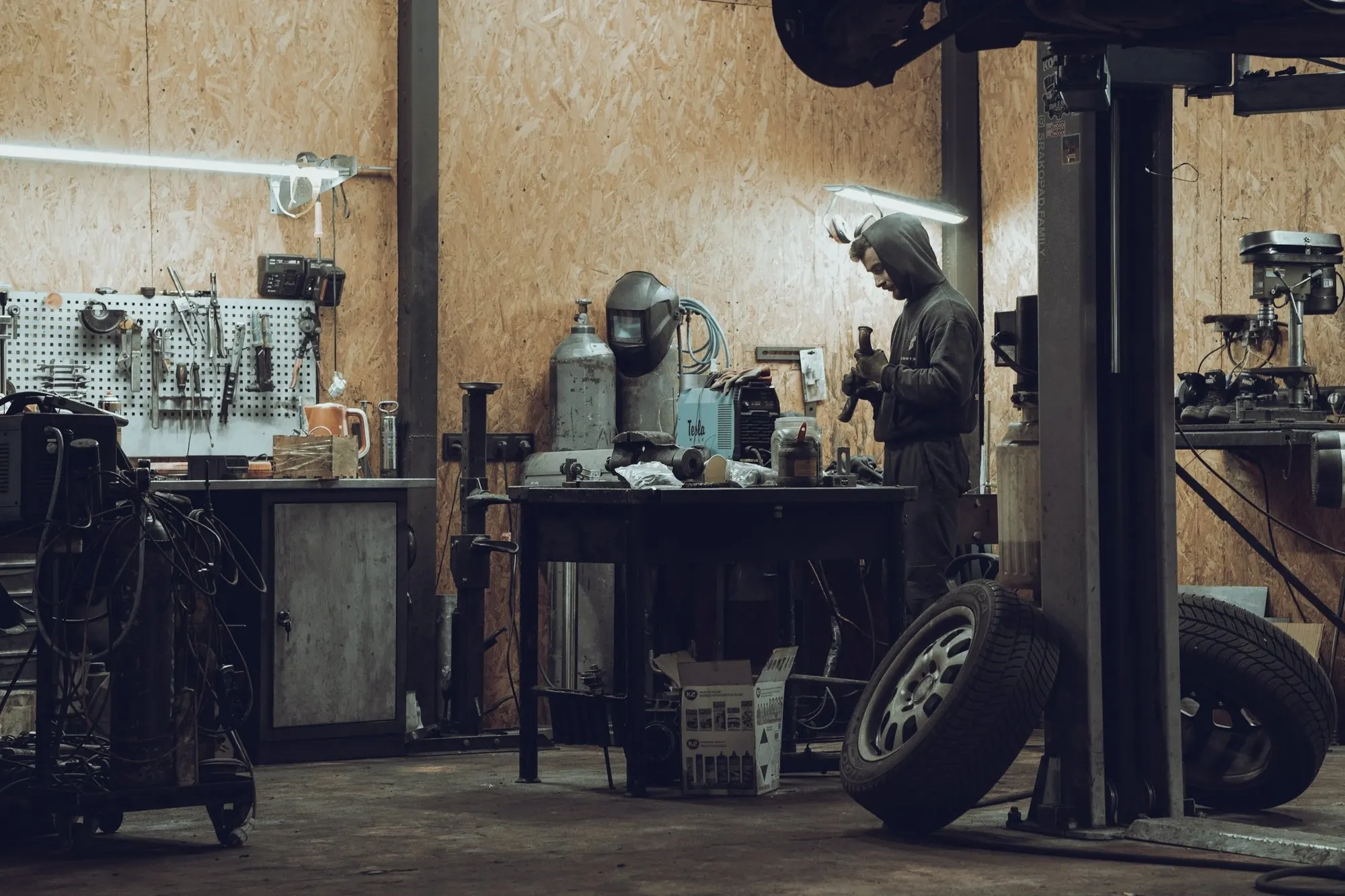 A mechanic works at a bench in a garage with a car lift and stacked tires.