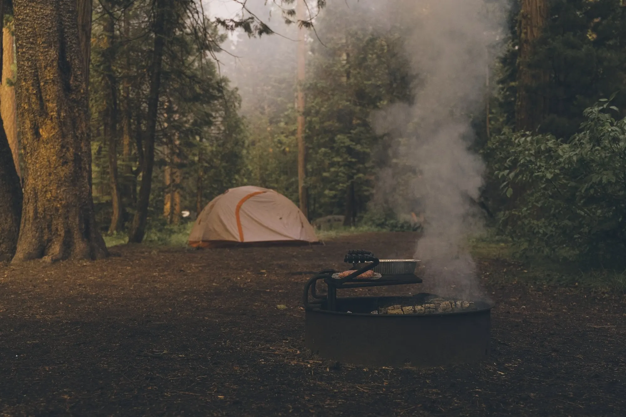 An orange tent pitched in a misty forest campsite beside a smoking fire ring.