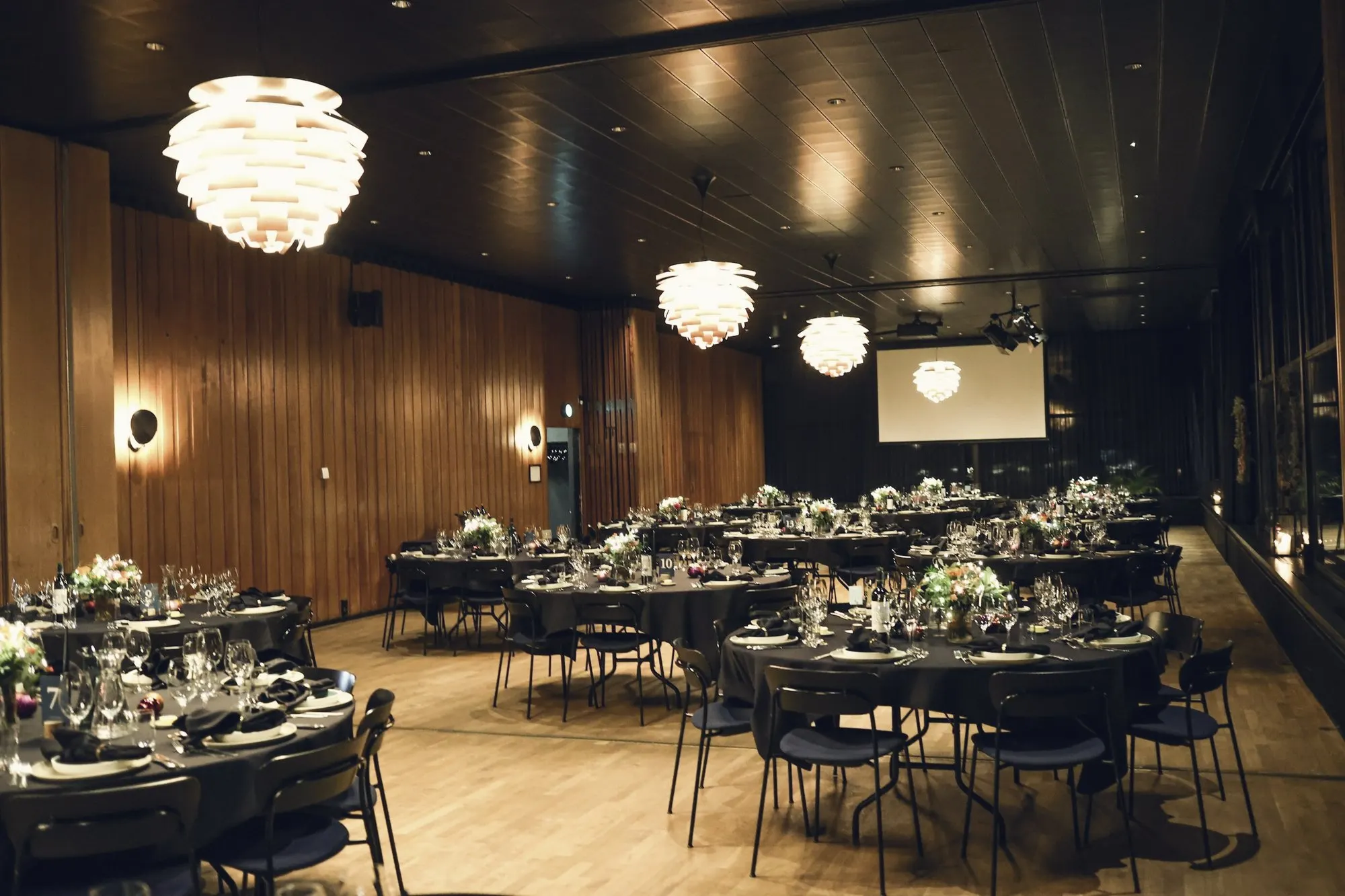 A wood-paneled banquet hall set with round tables under globe lights.