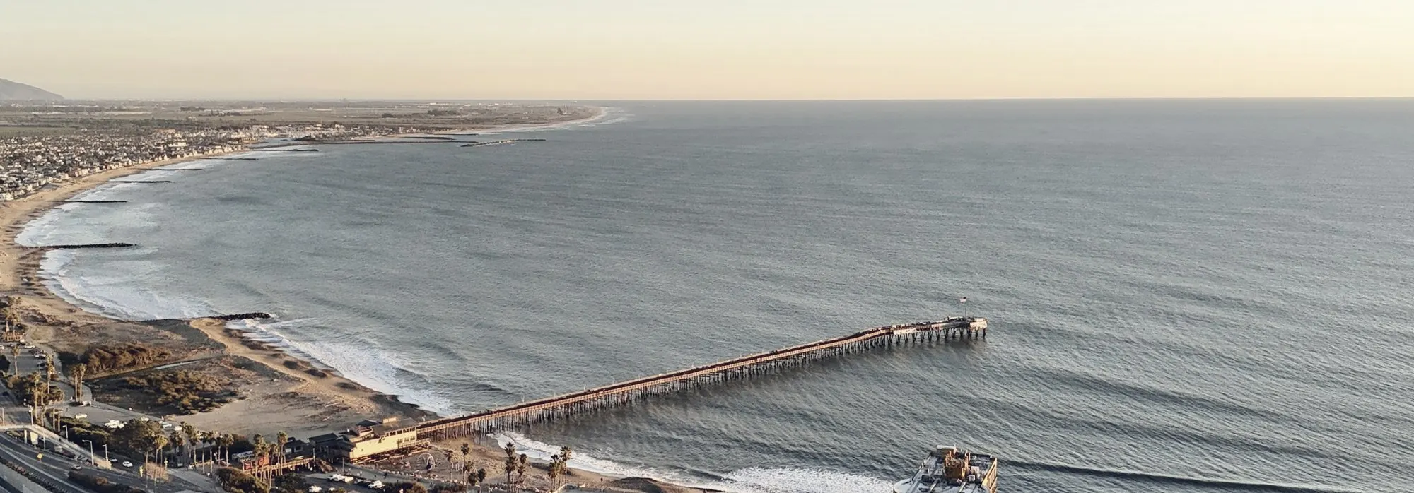 A long wooden pier reaching into the ocean along a curving Ventura County beach.