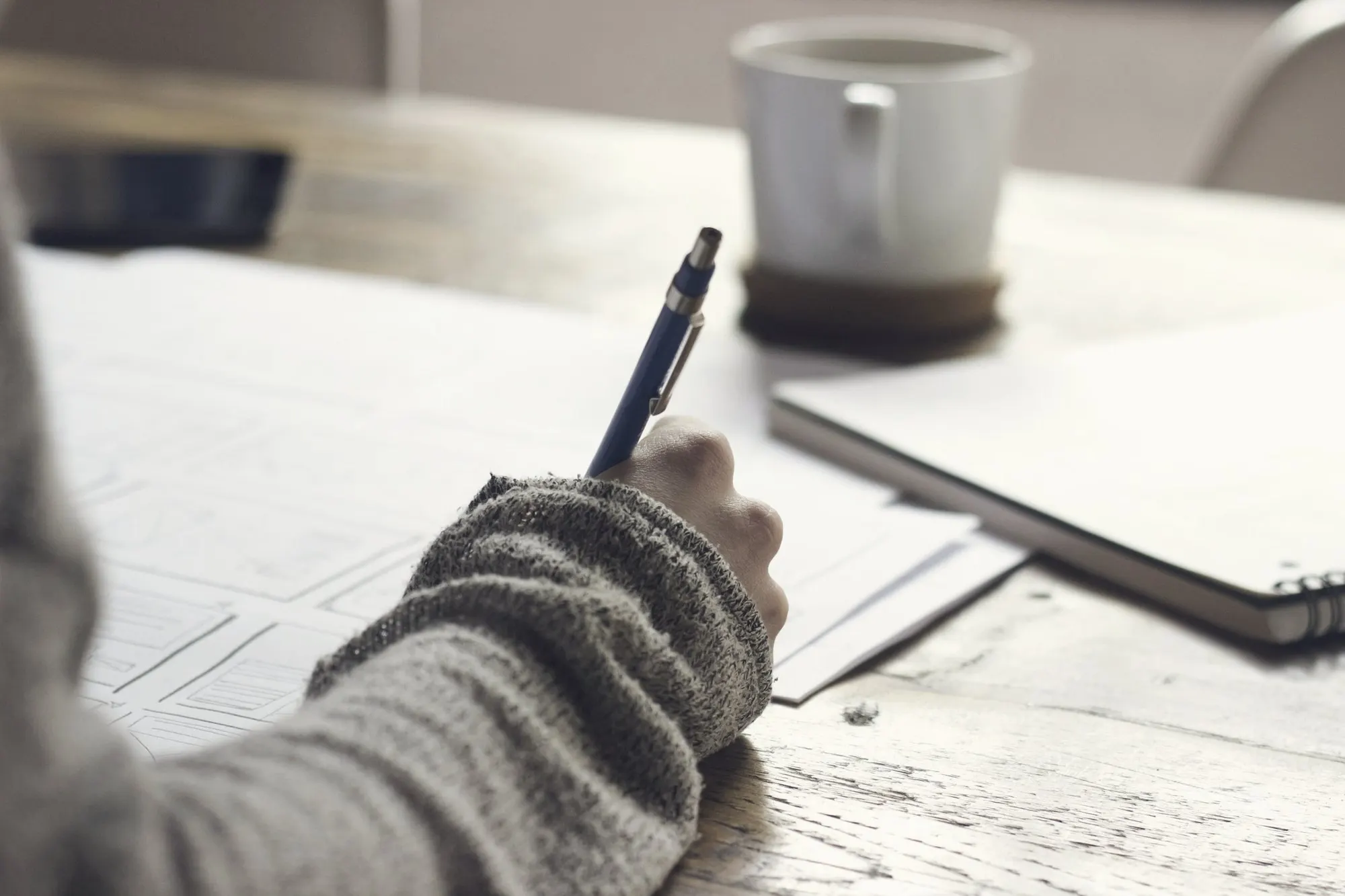 Hand writing on paper forms at a wooden desk beside a white coffee mug.