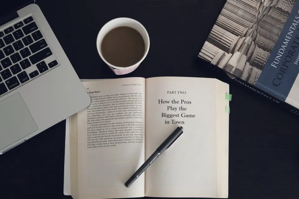 Open book, notepad, pen and coffee beside a laptop