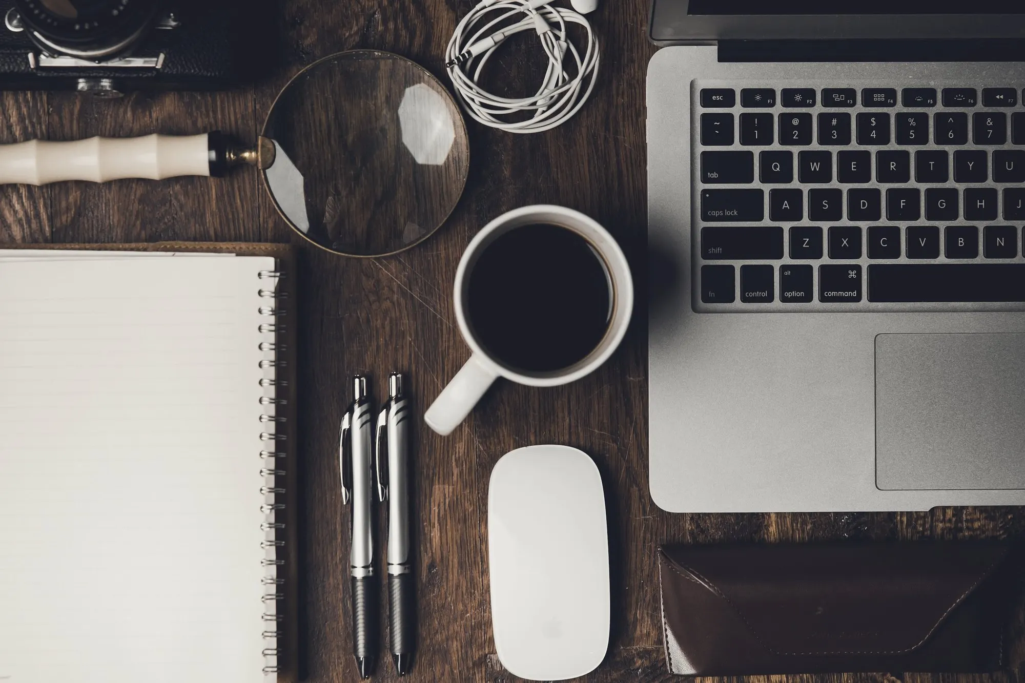 Wooden desk from above with notebook, pens, coffee cup, mouse and open laptop.