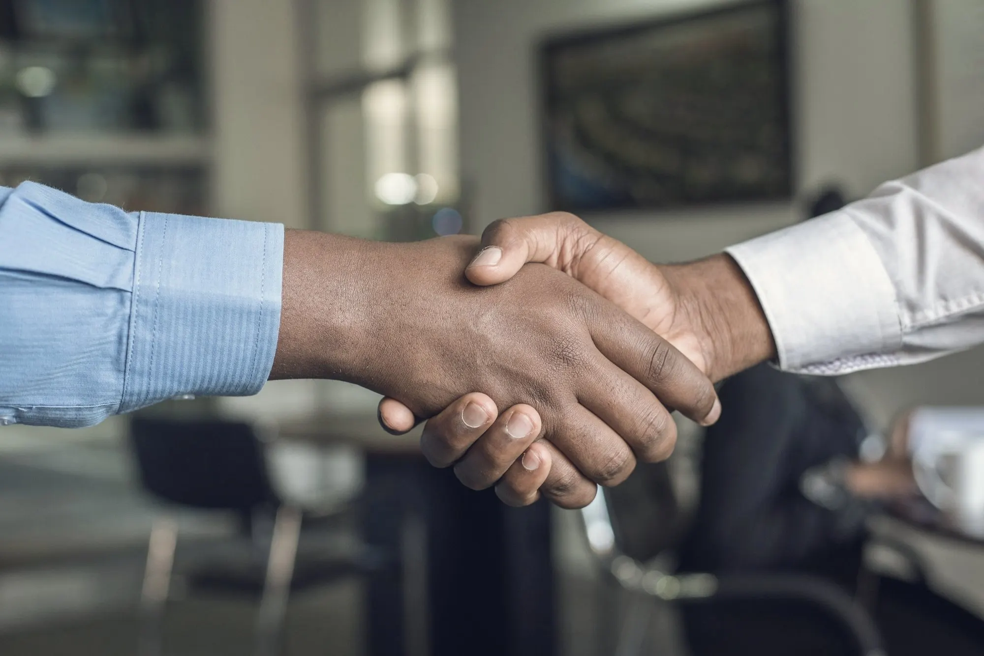 Two businesspeople in dress shirts shaking hands across a table in a modern office.