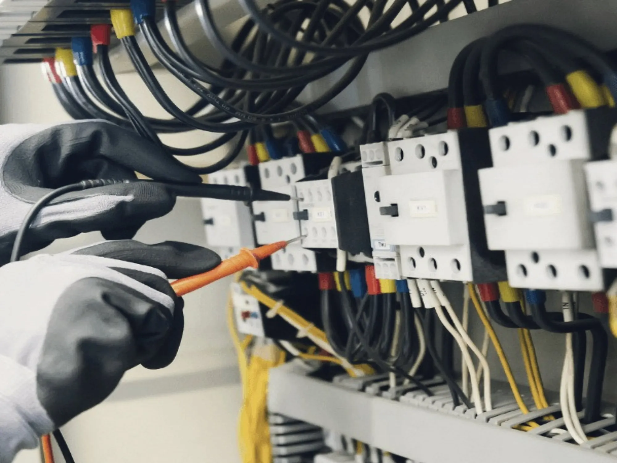 Gloved hands testing wired components inside an electrical panel with a multimeter probe.