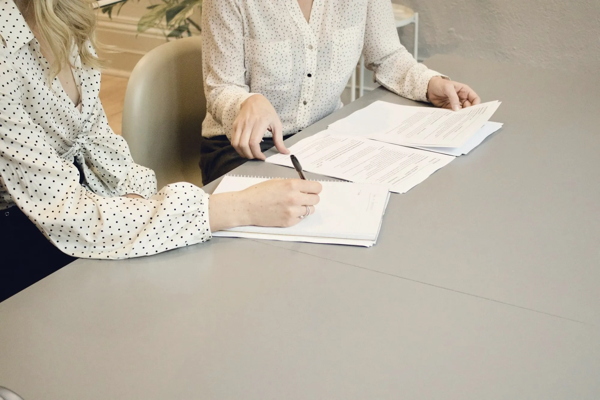 Two people at a grey table reviewing printed agreement pages, one writing notes with a pen.