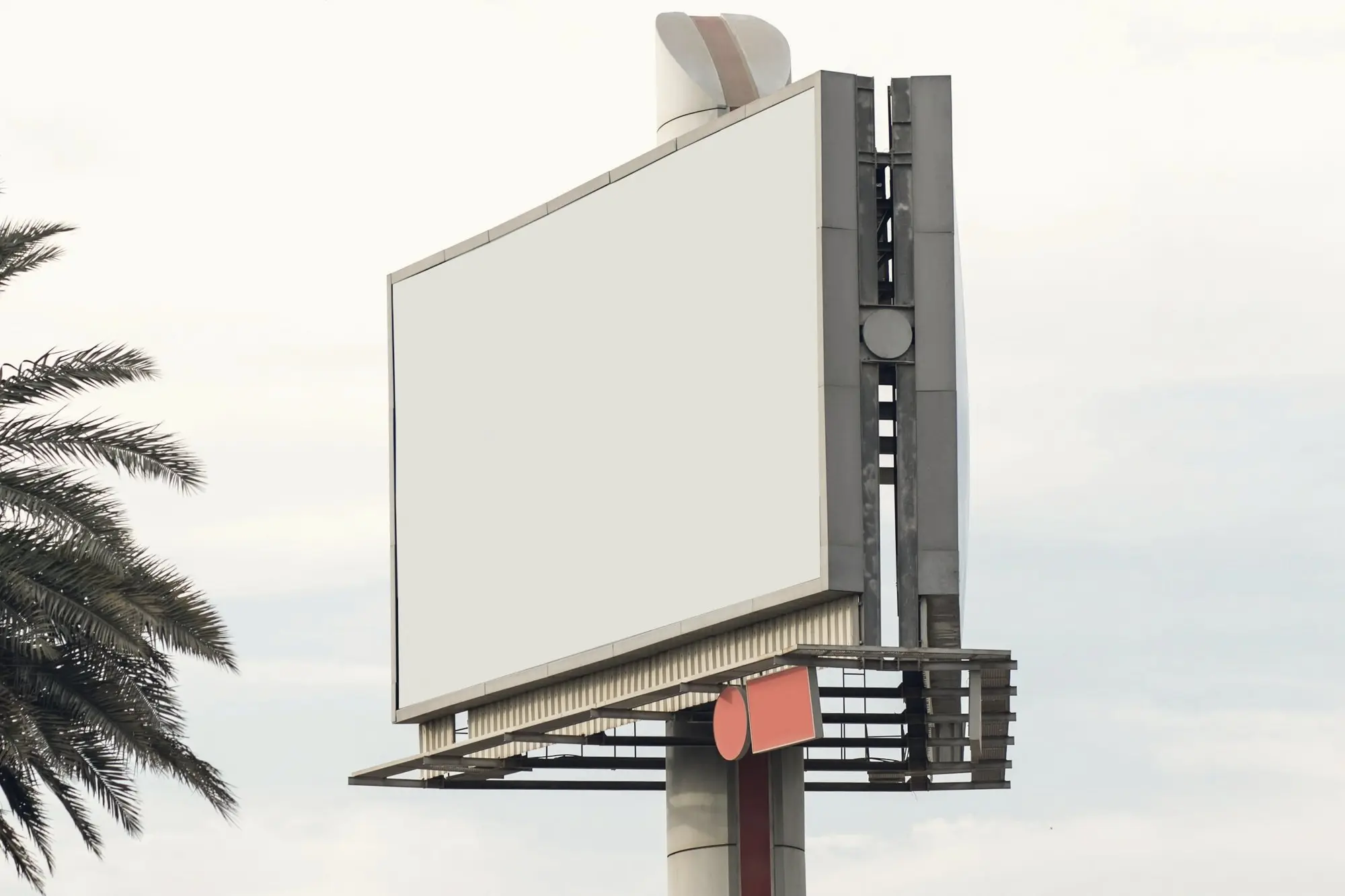 A blank billboard on a pole against a pale sky with palm fronds.