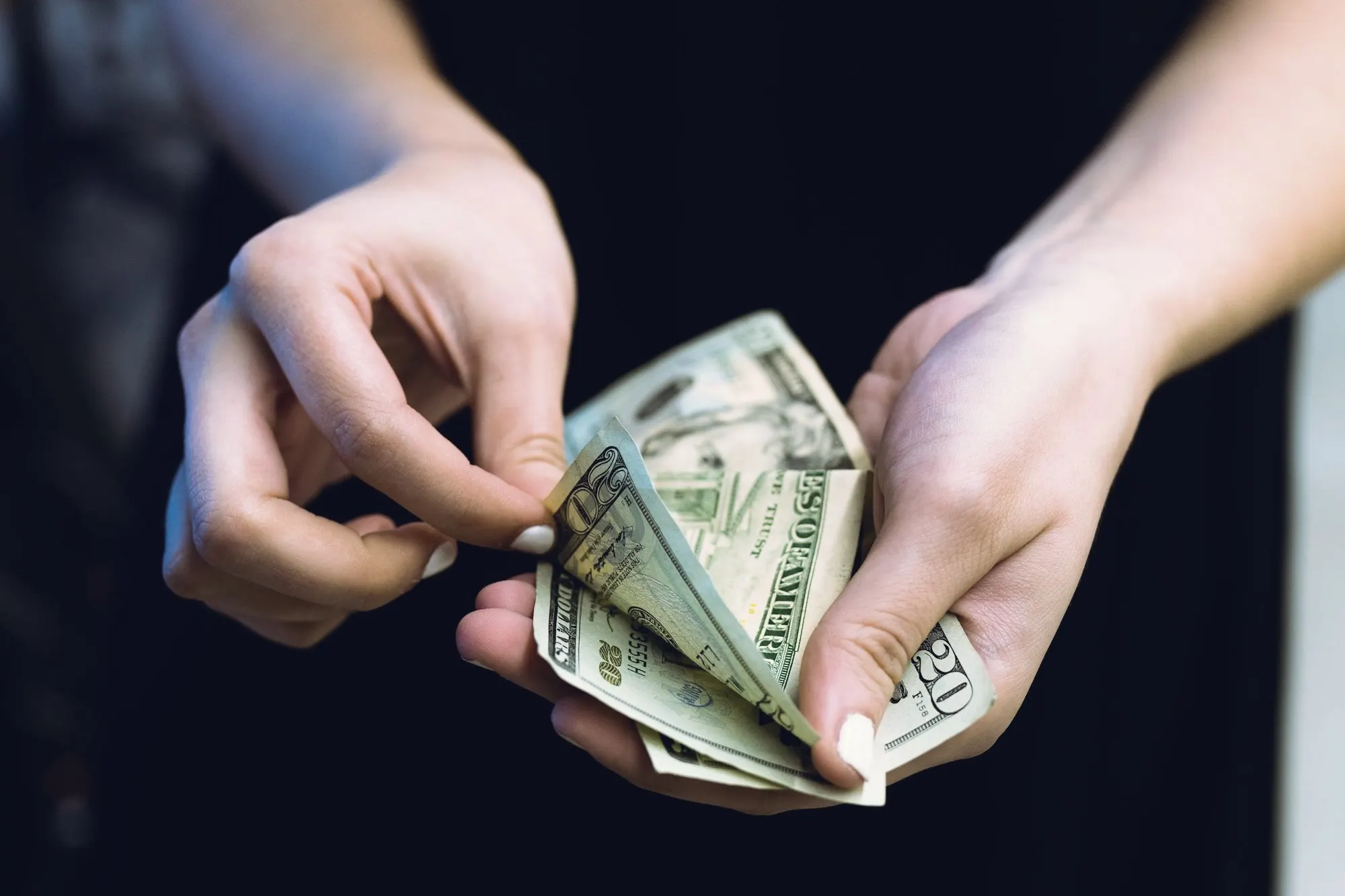 Hands counting a stack of dollar bills against a dark background.